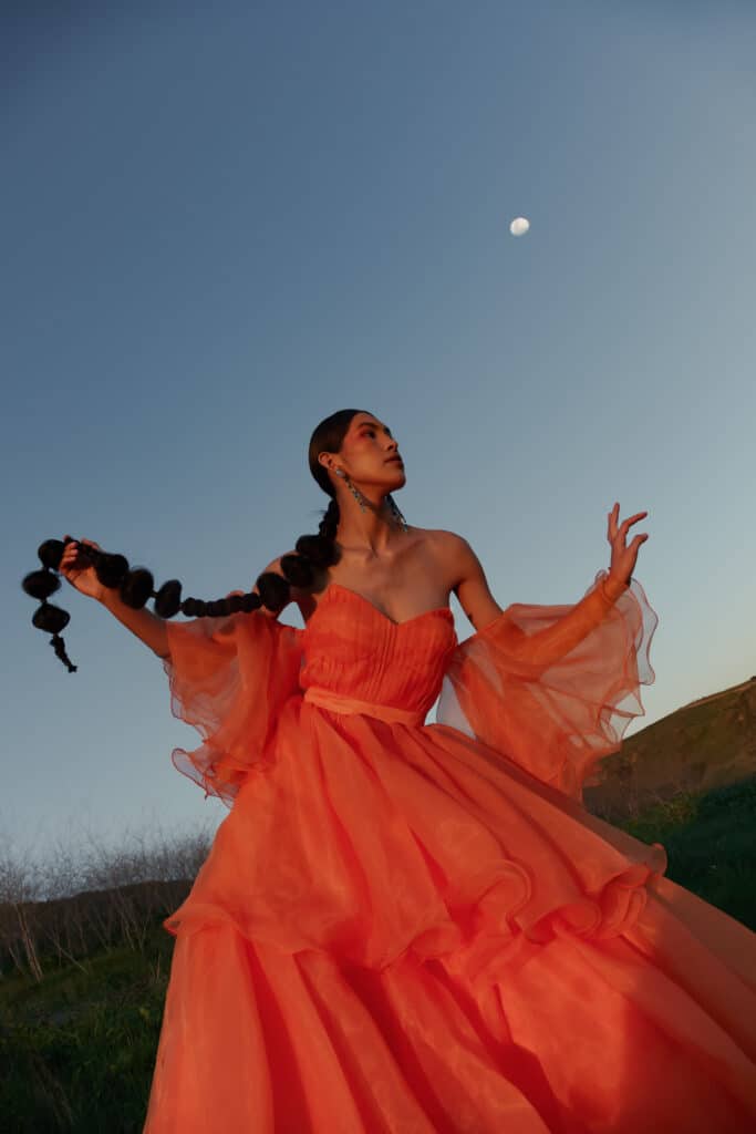 Woman wearing a poppy colored strapless, full-skirted gown posing holding her long braid.