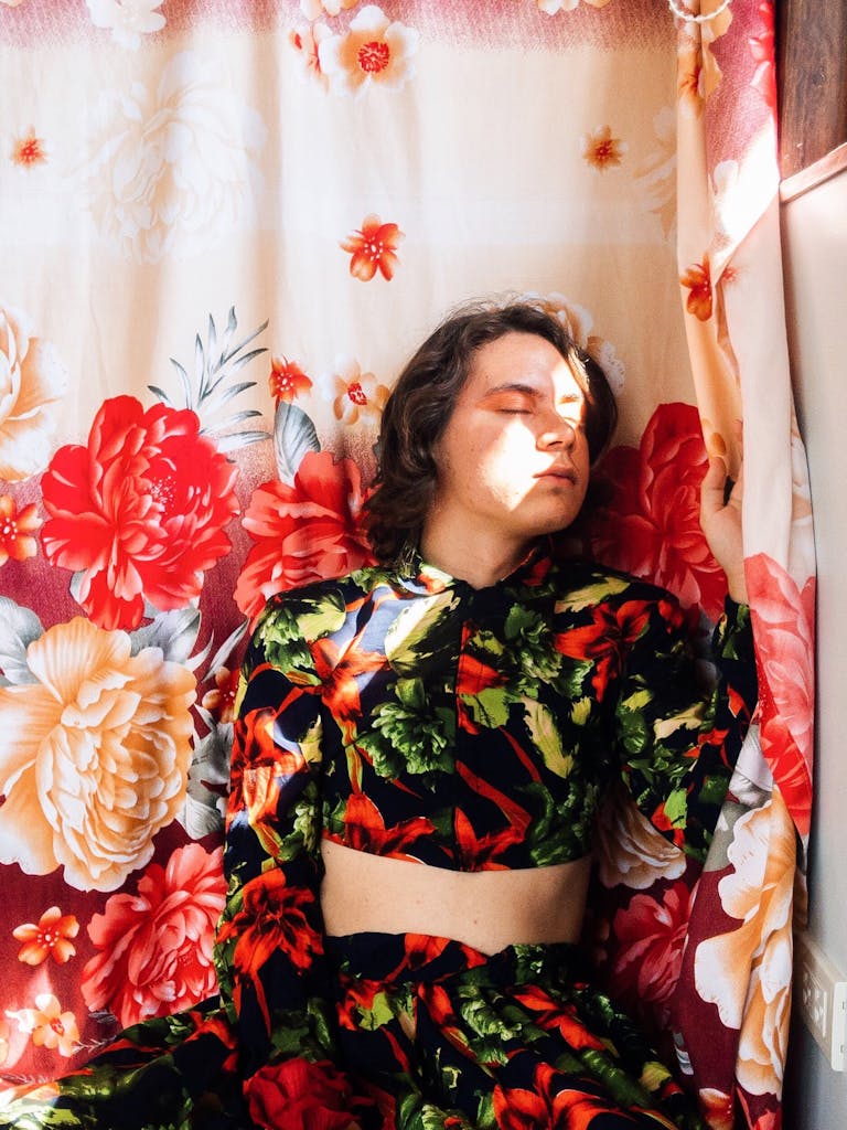 Person wearing floral dress with eyes closed facing a strip of light and an oversized floral textile hanging behind them