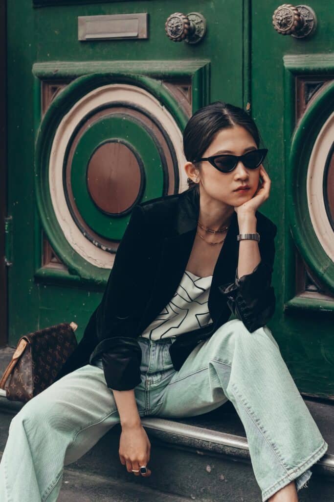 Woman wearing black sunglasses and blazer with light jeans sitting by a green door.